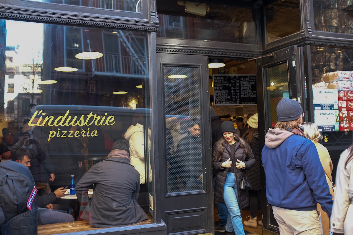 Busy L’Industrie Pizzeria storefront with customers inside and others queuing outside on a cold day.