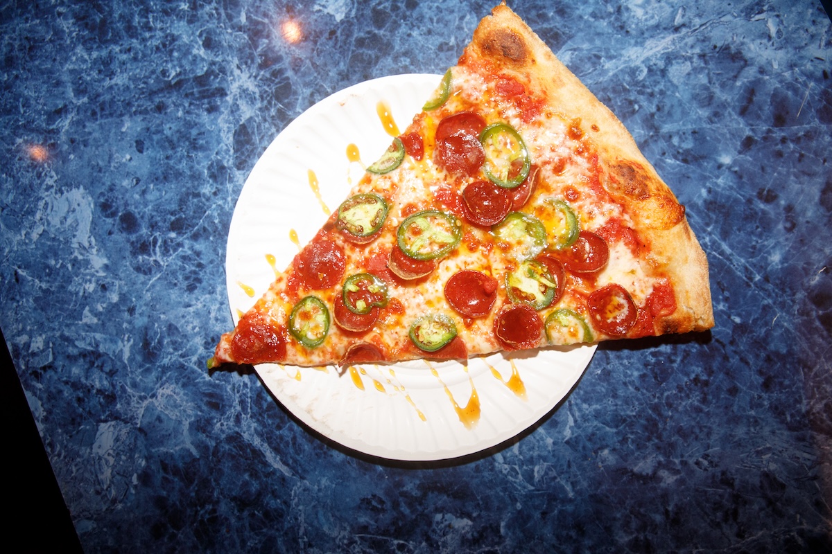 A single pepperoni and jalapeño pizza slice on a white plate atop a blue marble table.
