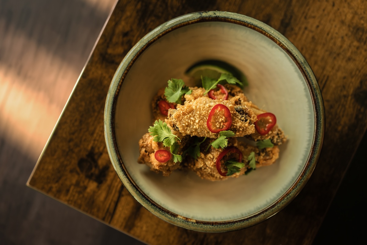 Crispy fried chicken bites topped with cilantro and sliced red chilies in a ceramic bowl on a wooden table with a lime wedge nearby.