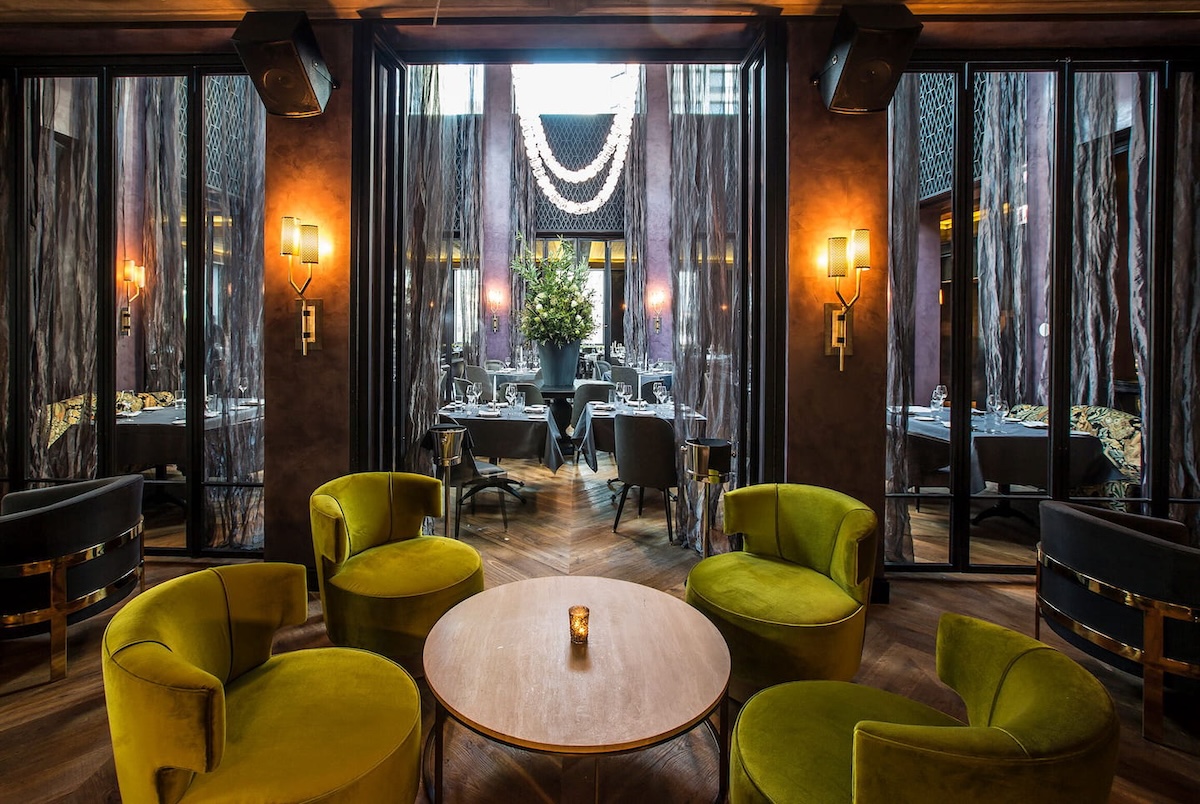 Luxurious restaurant lounge with lime-green velvet chairs around a round wooden table, candle centerpiece in warm lighting.