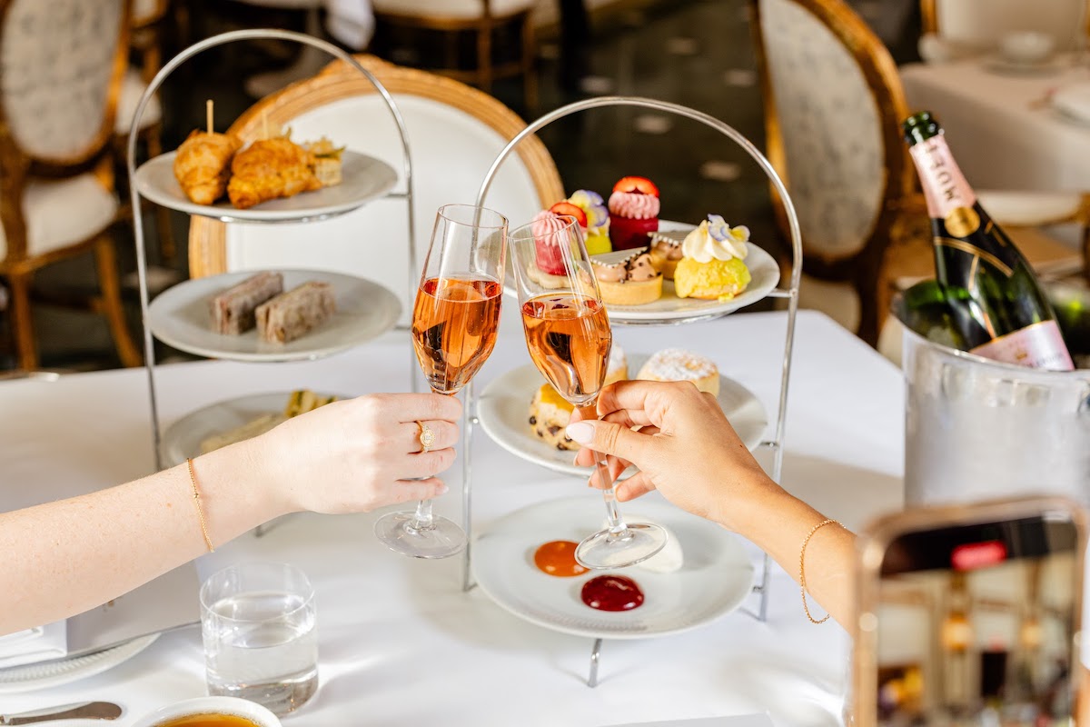 Two people toast with pink champagne flutes over a three-tiered dessert stand on a white table.