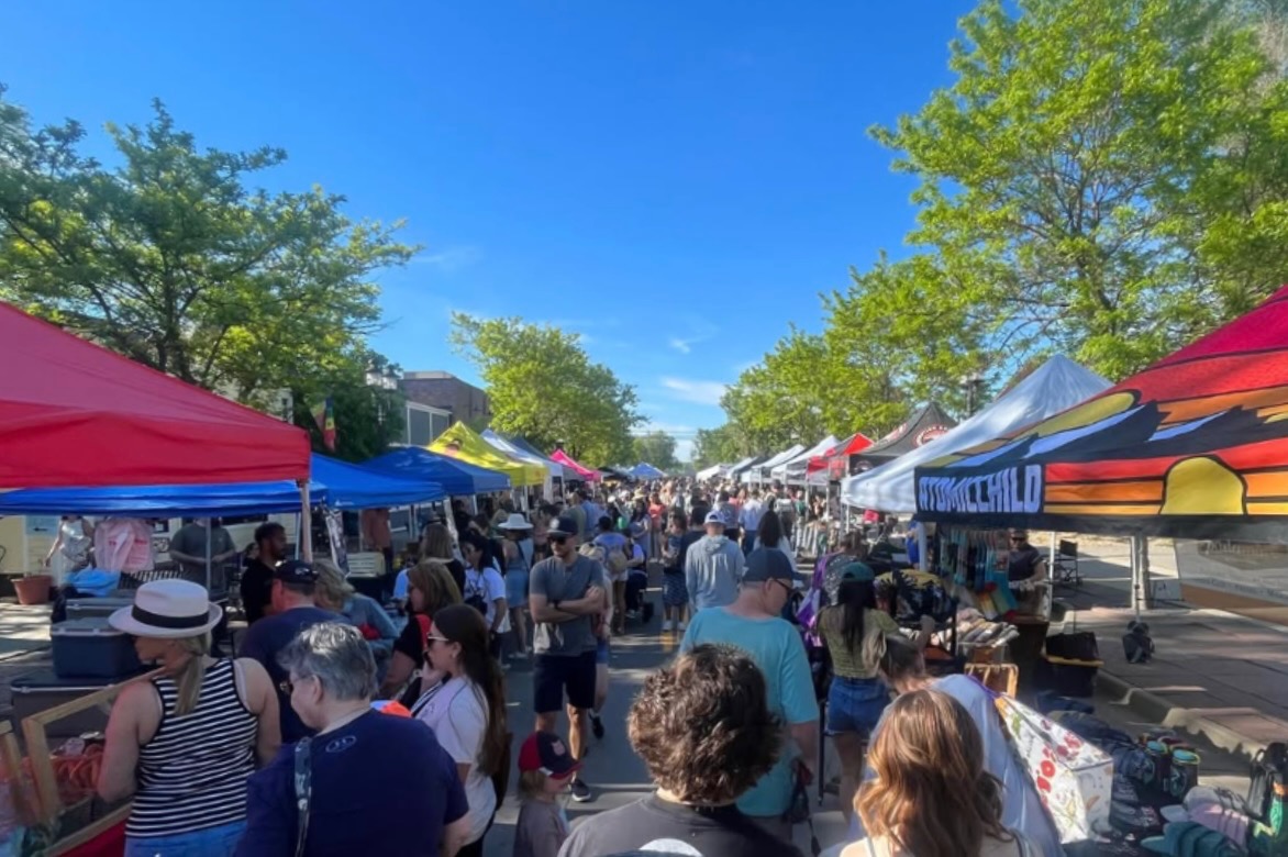 Busy outdoor street market with colorful tents and shoppers browsing stalls on a sunny day.