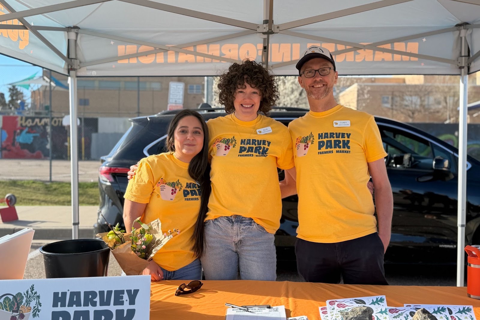 Three Harvey Park Farmers Market volunteers stand together under a canopy, smiling at the booth in yellow shirts with a car in the background.