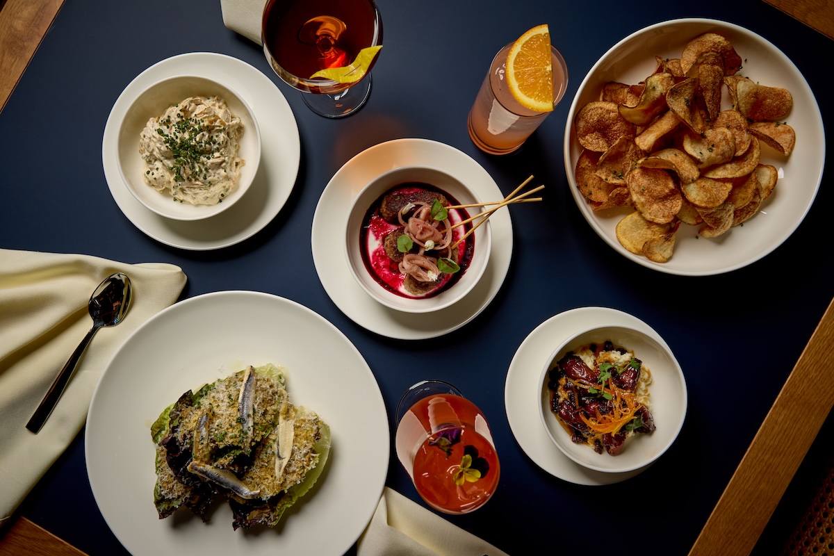 Top-down view of a dinner spread with potato chips, bowls of starters, and cocktails on a dark blue table cover.