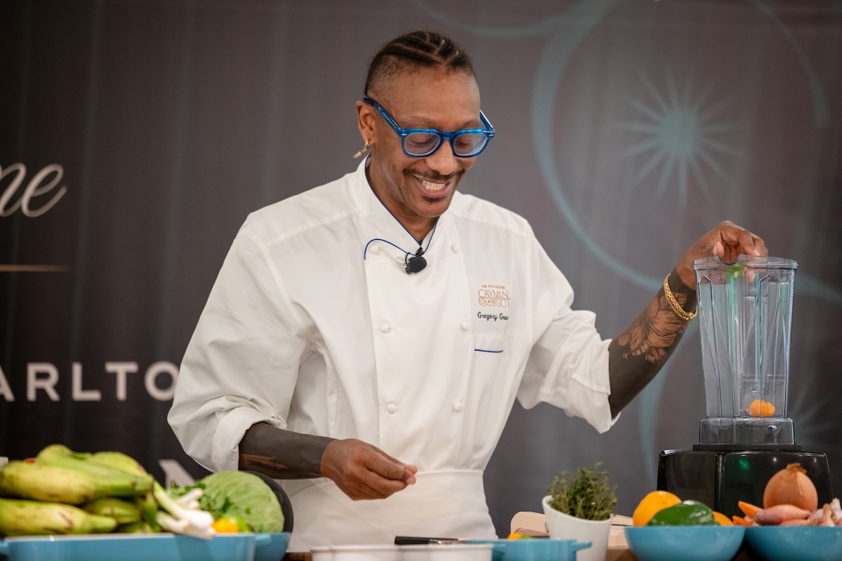 Chef Gregory Gourdet Demo_2 - DiningOut Chef in a white jacket and blue glasses demonstrates blending at a live cooking event, with fresh vegetables on the table.