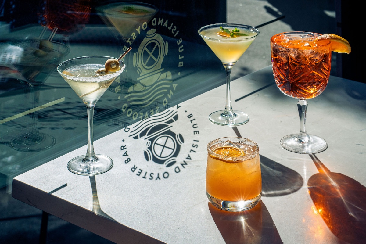 Five drinks on a sunlit white bar table: two martinis with olives, a pale yellow cocktail, an orange short drink with a citrus garnish, and a red cocktail with an orange slice.