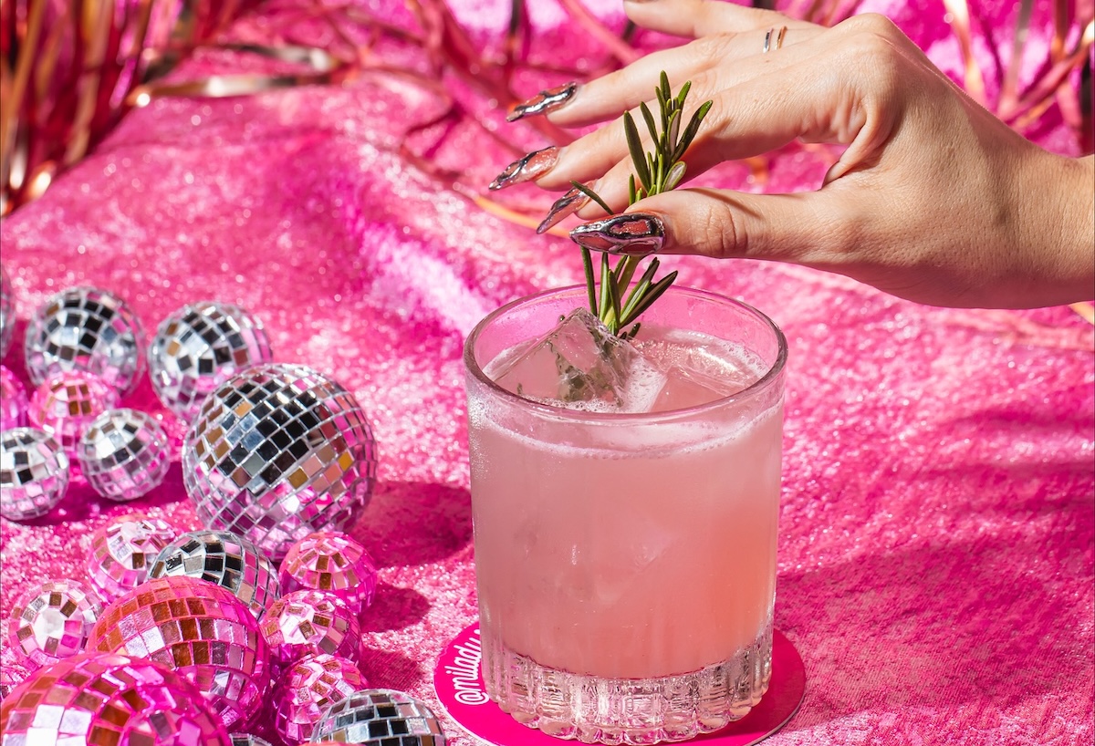Pink cocktail in a short glaWhy simply wait when you can go drink before your table is ready. | Photo courtesy of Milady'sss on a glittery pink surface, rosemary garnish held by a hand with metallic nails.