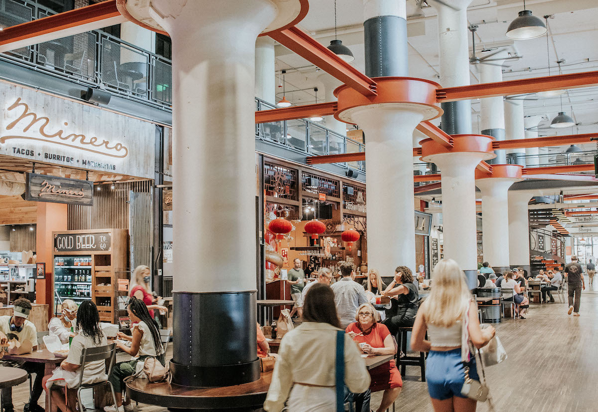 Indoor food hall with large white columns and orange beams; diners sit at tables near a Minero taco shop sign.