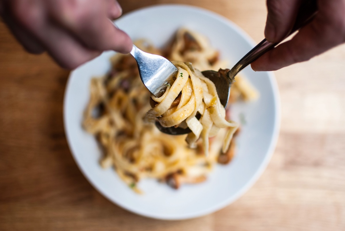 Two forks lift and twirl creamy fettuccine pasta with mushrooms from a white plate on a wooden table.