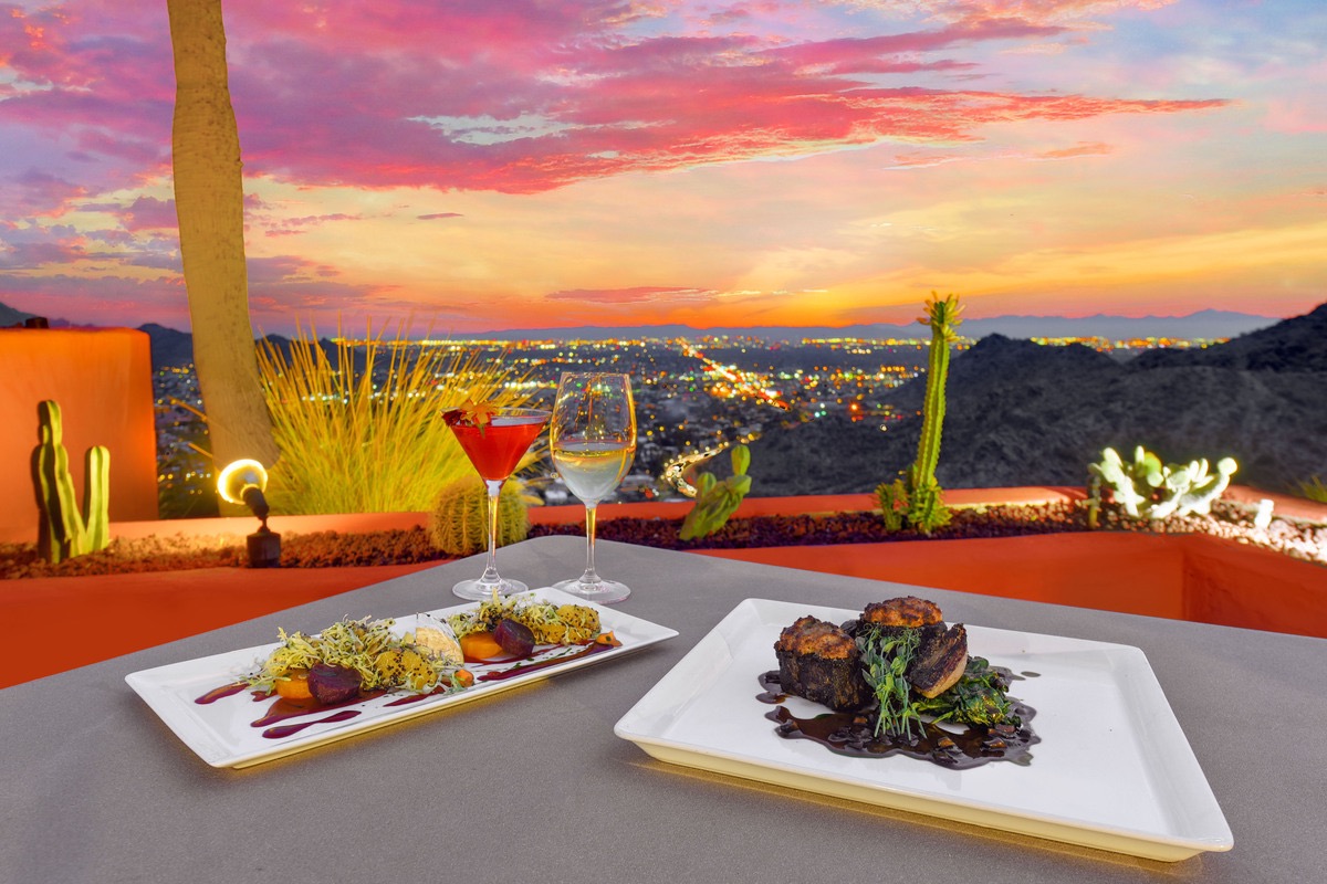 Outdoor sunset dinner on a terrace with two plated dishes, two glasses, and a colorful cityscape below.