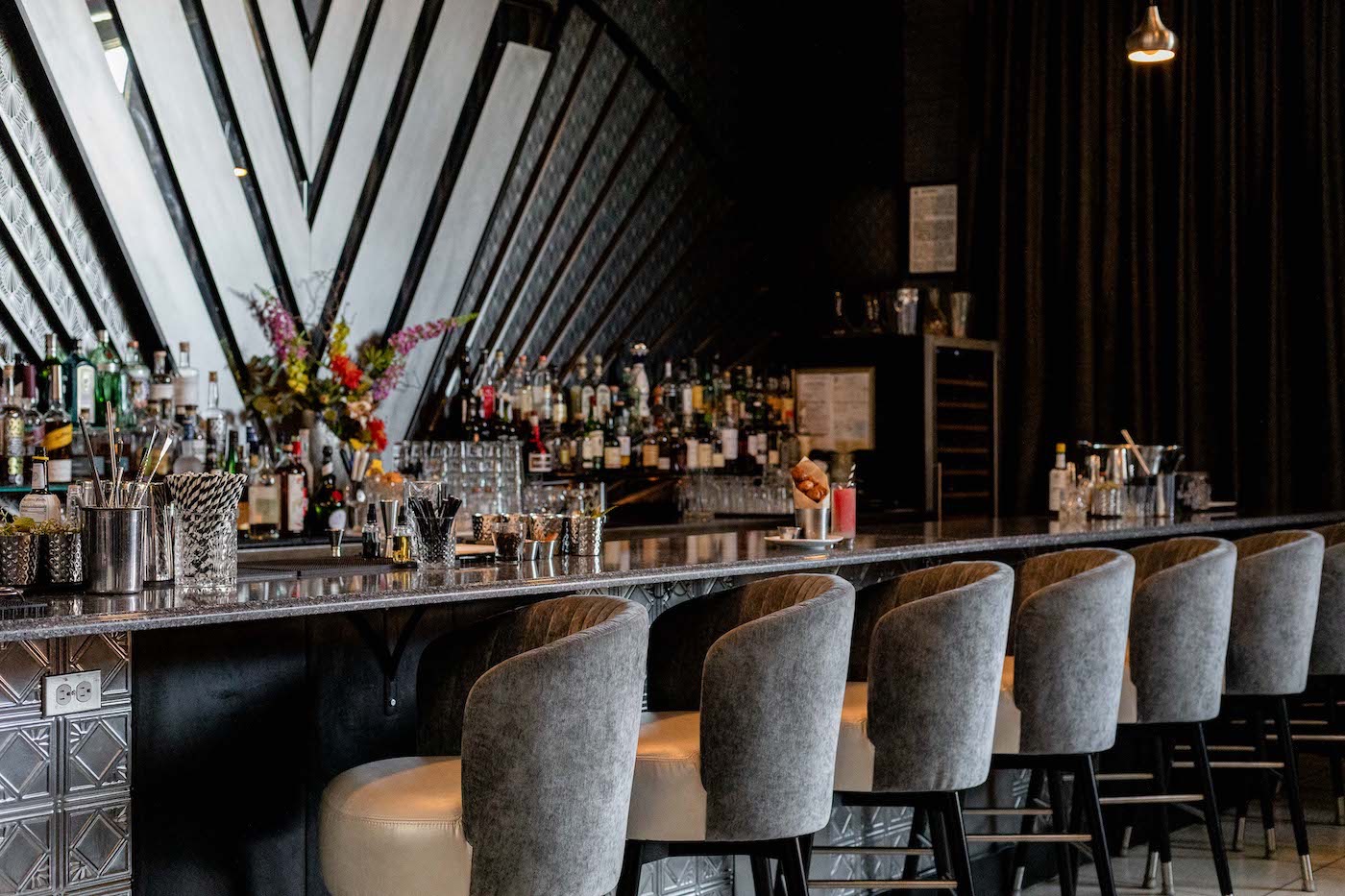 Long bar with a marble counter, gray upholstered stools, and bottles lined up behind the bar.
