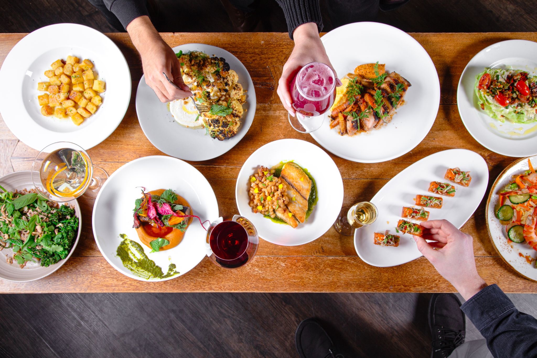 Top-down view of a long wooden table with multiple white plates of varied dishes, drinks, and hands reaching for food beneath a dark-sleeved sleeve.
