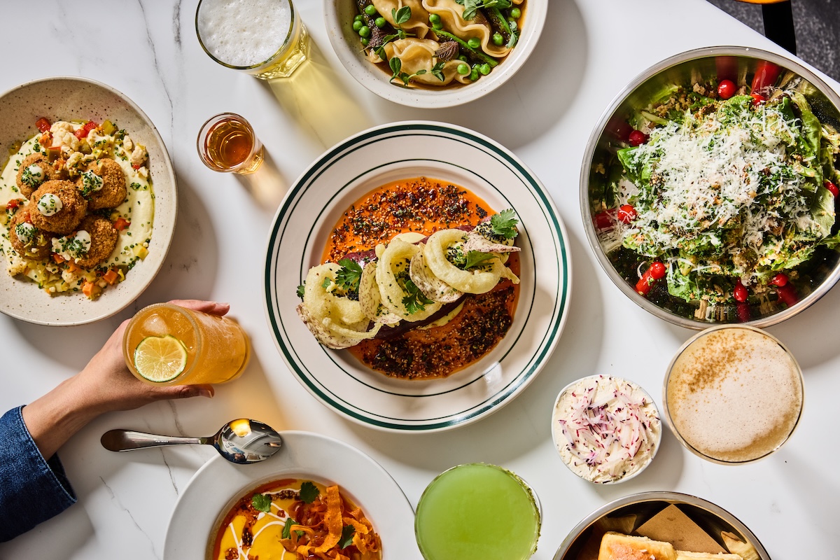 Top‑down view of a shared feast: central plate with meat in red sauce and onion rings, surrounded by salads, croquettes, pasta, and drinks.