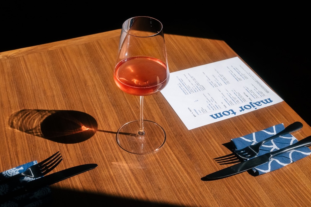 Wine glaHead to Major Tom in RiNo for great food and wine. | Photo by Joe Freemond denver ss with pink wine on a wooden table, shadow cast, near a menu and blue patterned napkin with forks and knife nearby.