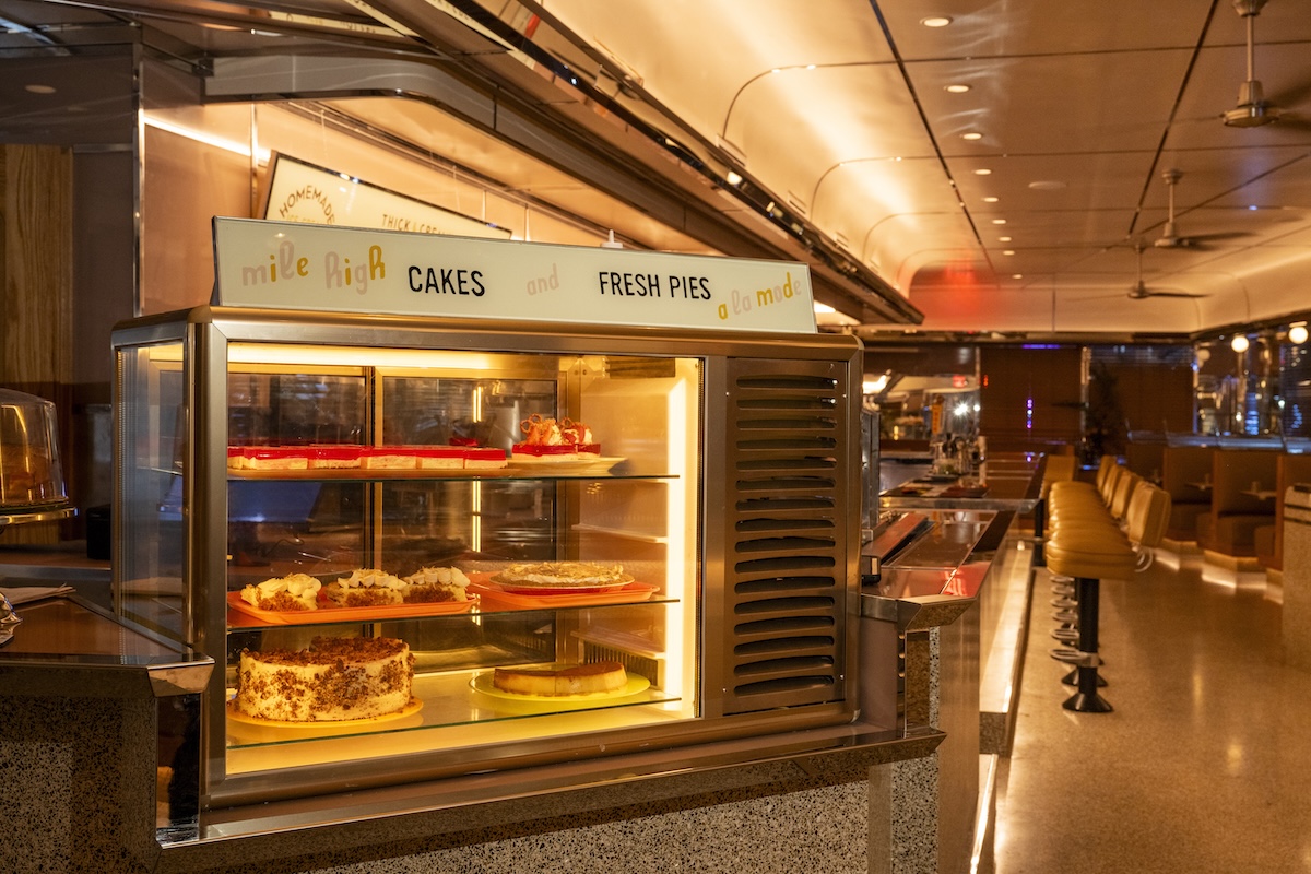 NYC_Evan Sung_Kelloggs_5 - DiningOut Retro diner bakery display casKellogg’s Diner offers comfort both in food and setting. | Photo by Evan Sunge with cakes and pies under warm lighting; sign says'mile high CAKES and FRESH PIES'.