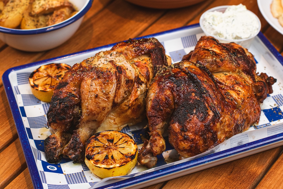 Two roasted chickens with charred skin on a blue-and-white patterned tray, with grilled lemon halves and a small herb dip on the side.