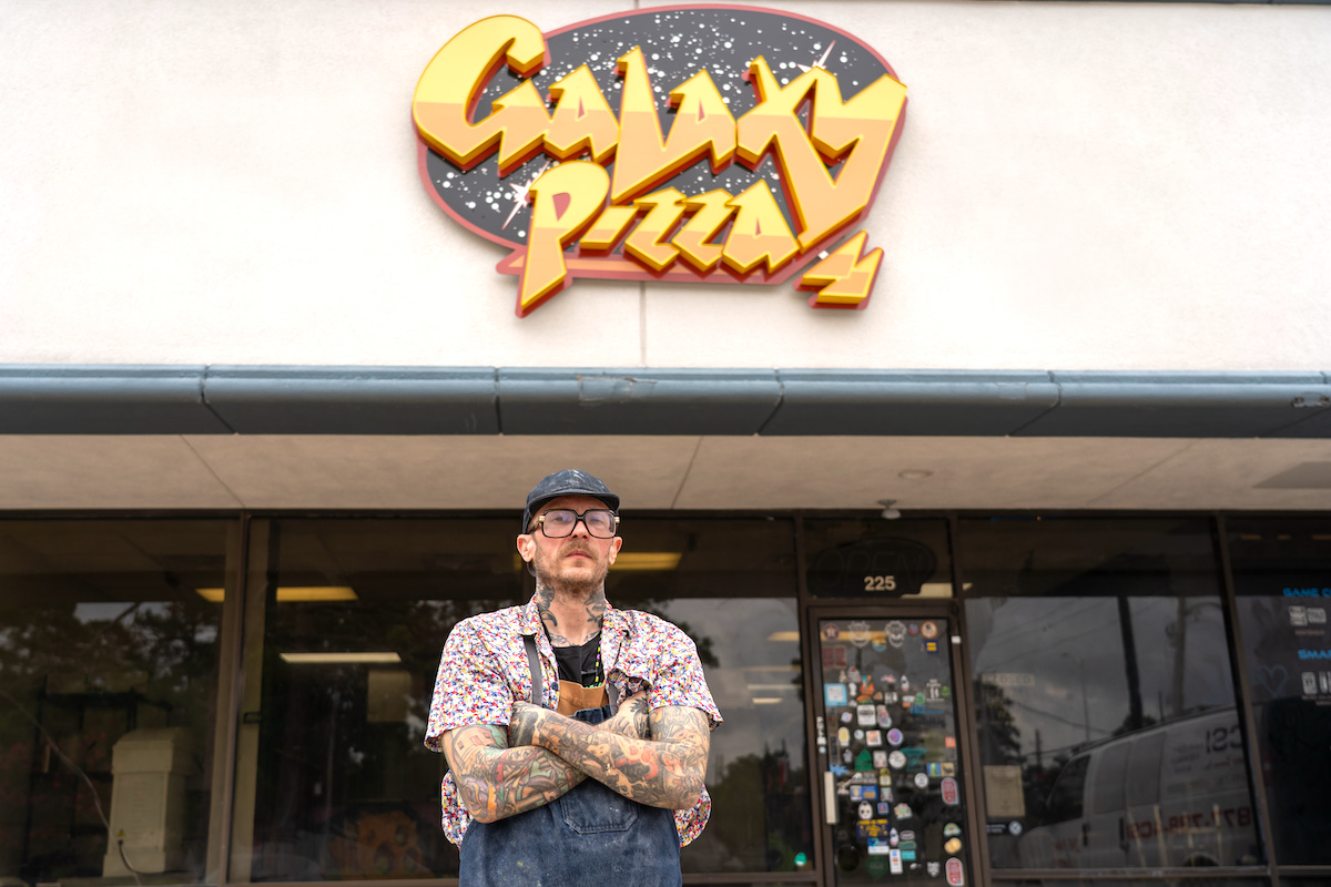 Tattooed man in a denim apron stands with arms crossed in front of Galaxy Pizza storefront sign.