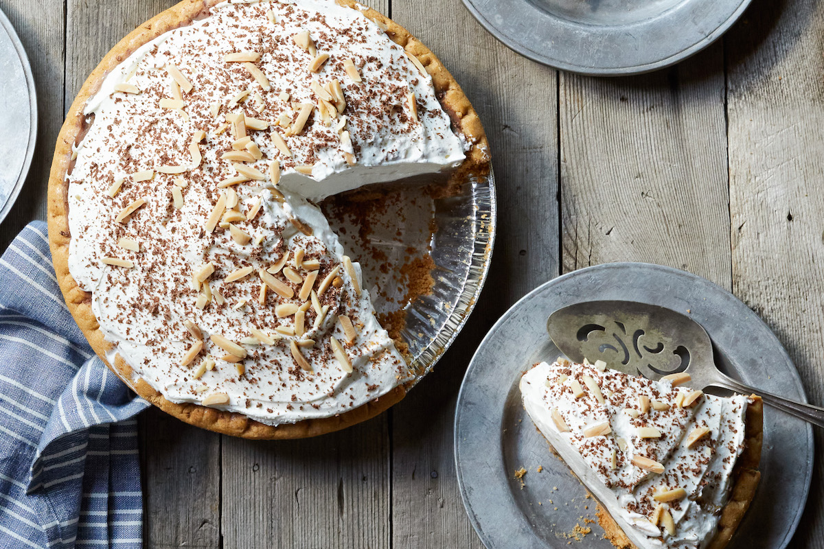 Whole cream pie topped with whipped cream, chocolate shavings and almond slivers on a rustic wooden table; a slice has been removed from the pan.