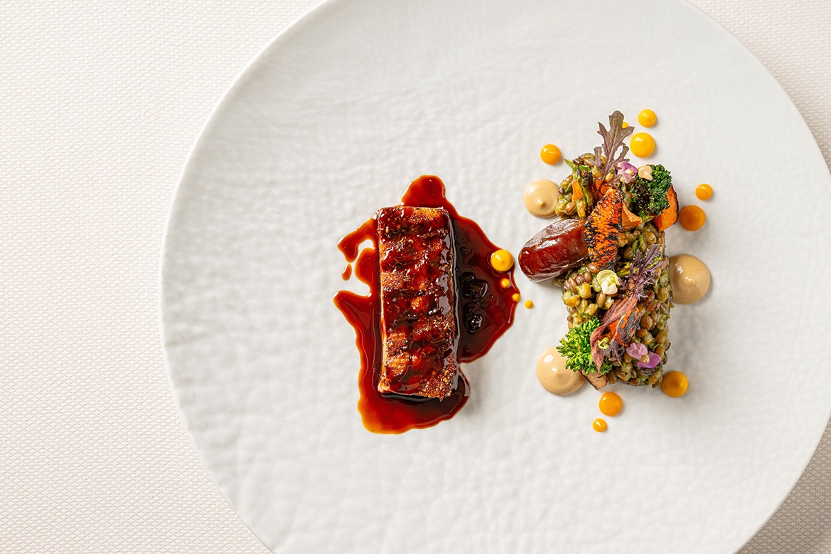 Plated gourmet dish with a glazed meat slice in red sauce and a colorful grain-and-vegetable cluster on the side, drizzled sauces around the plate.
