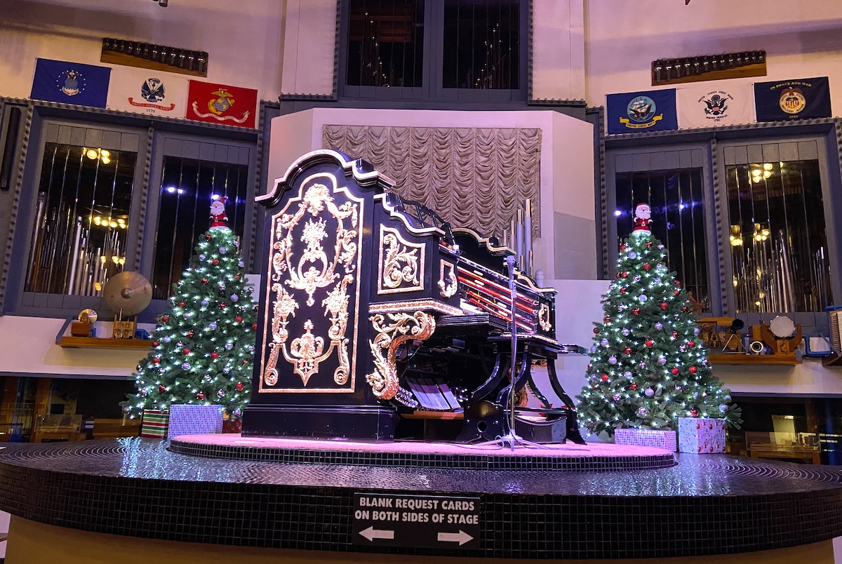 Ornate black and gold pipe organ on a circular stage, flanked by lit Christmas trees and wrapped presents.