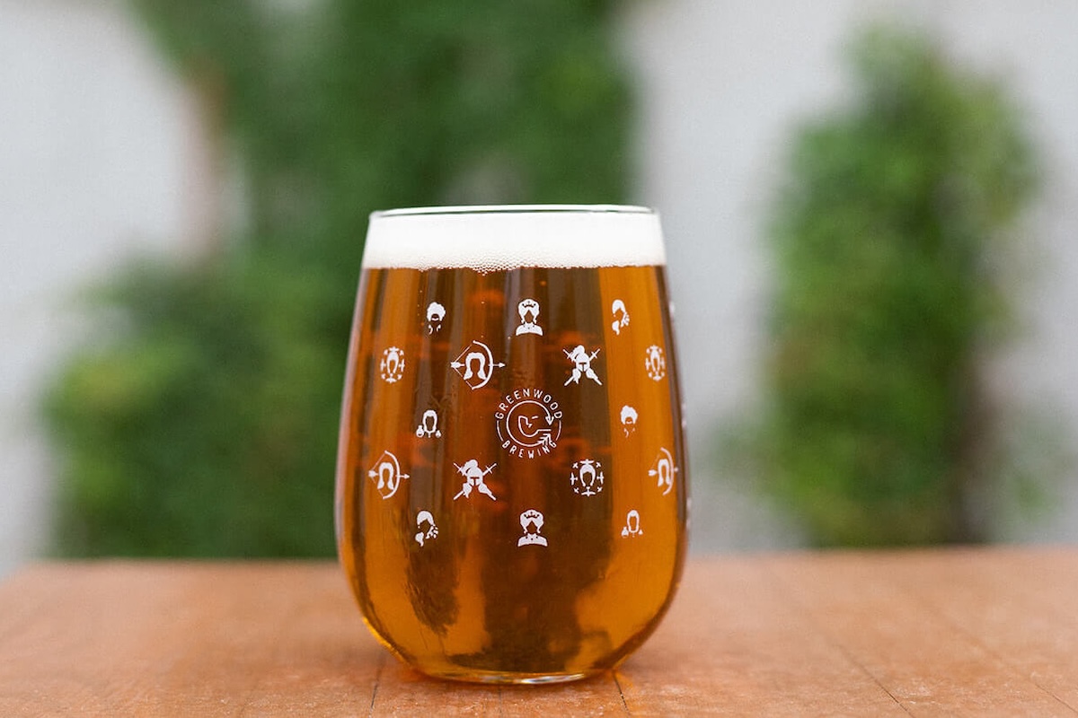 Amber beer in a stemless glass with a foamy white head on a wooden table, outdoors with greenery in the background.