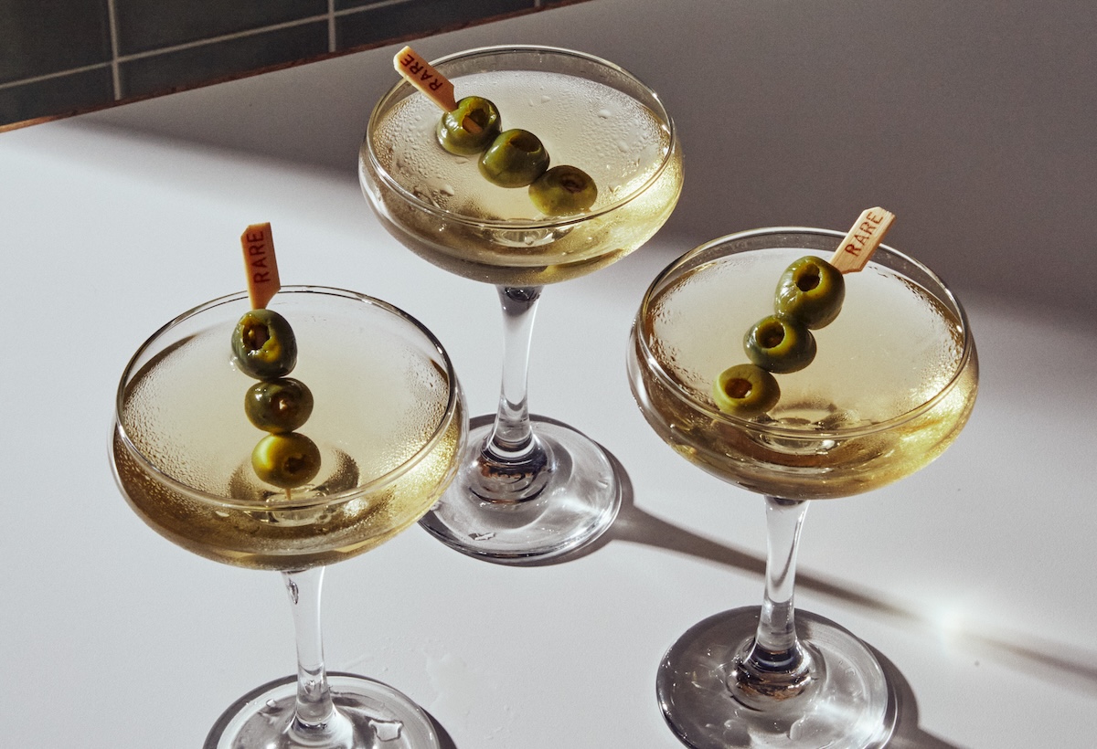 Three coupe glasses of pale cocktail, each with olive skewers labeled RARE resting on a white surface.