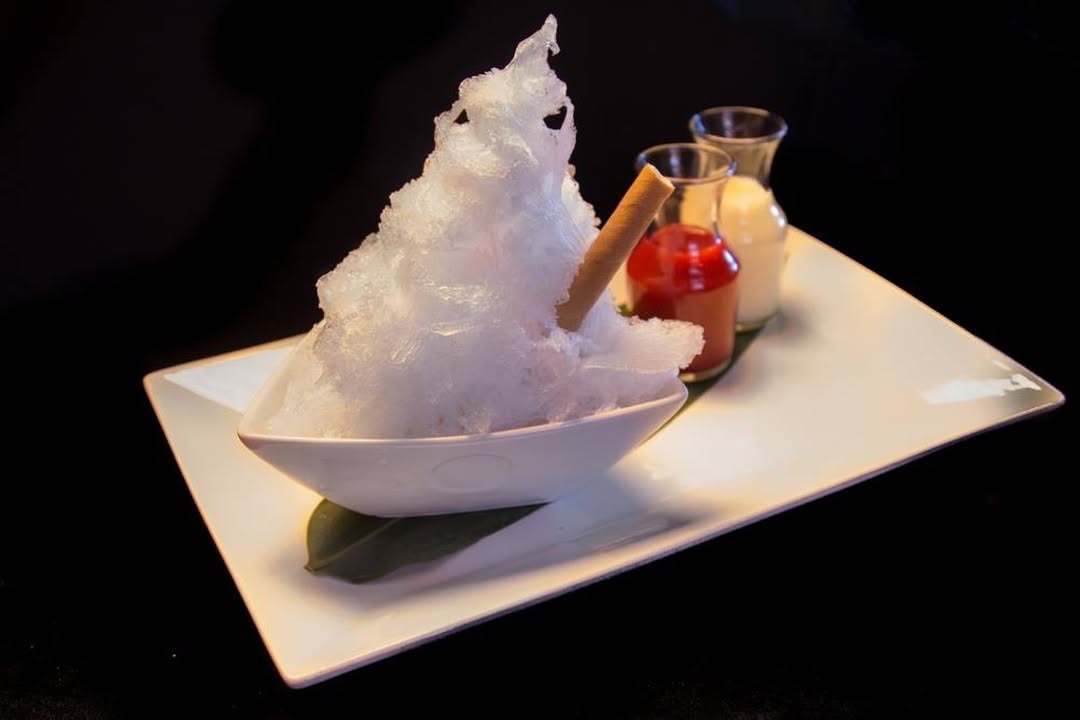 The flavor of Matsuhisa’s kakigōri changes seasonally | Photo by Matsuhisa - DiningOut White bowl of shaved ice piled high with a cinnamon stick, on a rectangular plate with small sauce jars in the background.