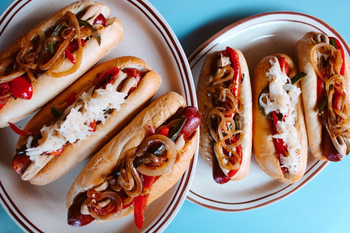 Two hot dogs topped with onions, peppers, and sauerkraut on white plates against a blue background for a casual meal setting.