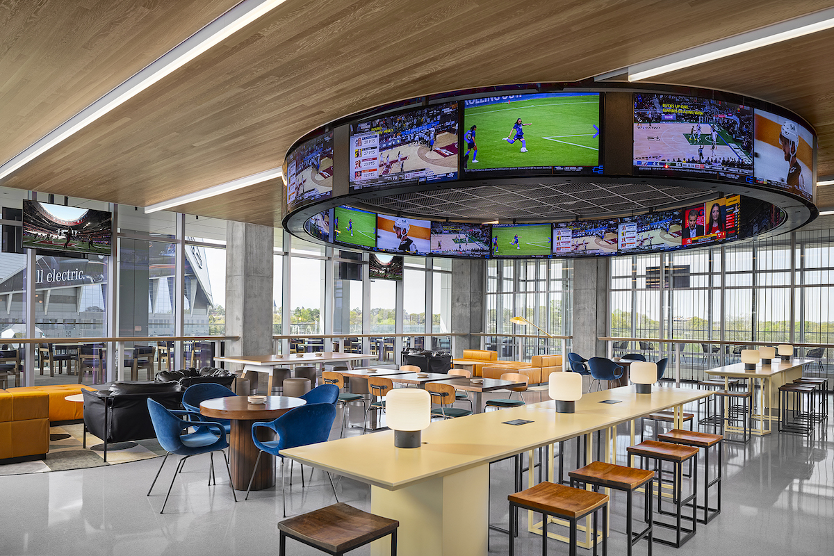 Open, modern lounge area with a circular ring of TV screens overhead showing sports, surrounded by blue chairs, beige high tables, and large windows.
