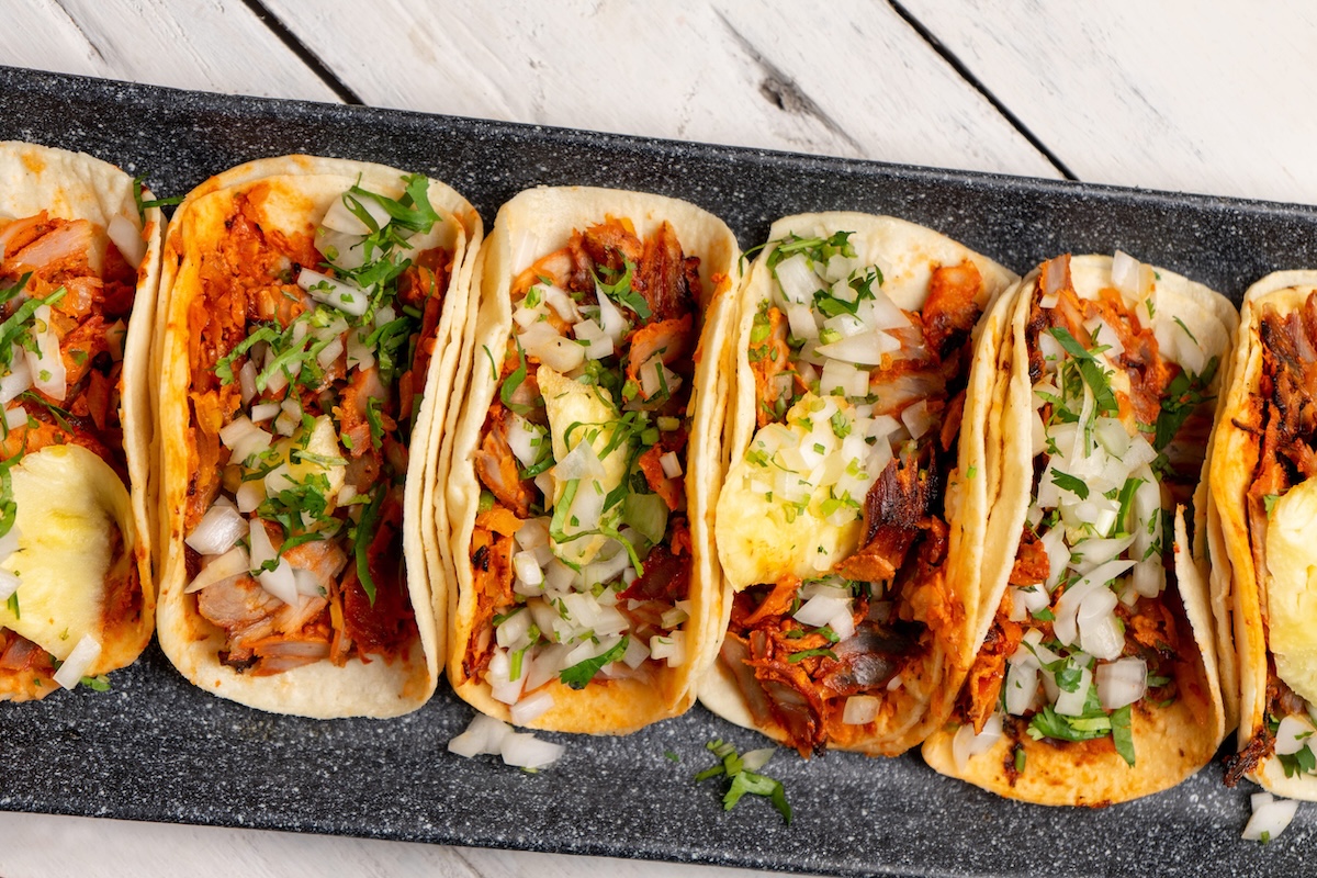 Row of soft tortillas filled with shredded meat, onions, cilantro, and cheese on a dark tray. Traditional mexican pork tacos called"Al pastor" with pineapple on white background