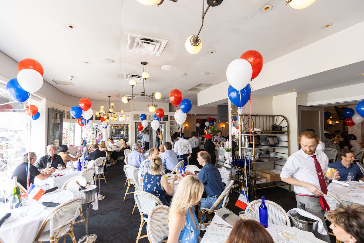 Bustling restaurant during a celebration; white tables with blue bottles, red‑white‑blue balloons, and flag decorations.