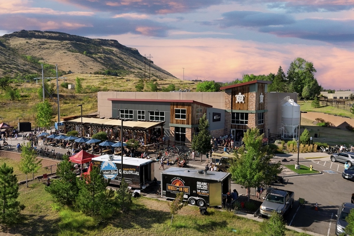 Aerial view of a busy outdoor market and café in front of a modern building, with food trucks and parked cars nearby.
