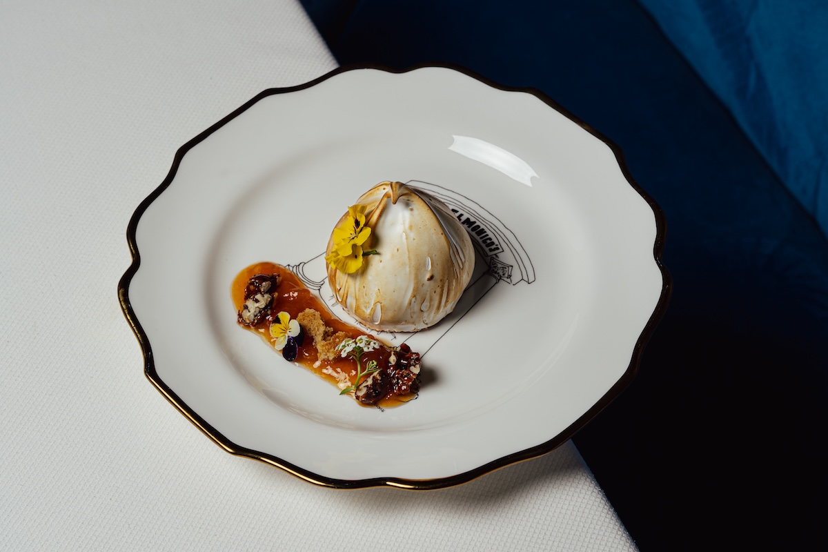 NYC_Delmonicos_baked_alaska - DiningOut Elegant plated dessert: a white domed pastry with a yellow edible flower, paired with caramel sauce and toppings on a white, gold-rimmed plate.