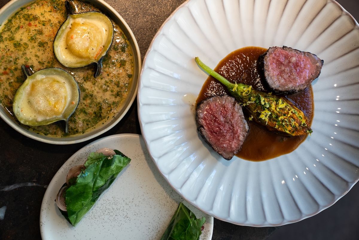 NY_Cynthias - DiningOut Overhead view of a fine-dining plate: two slices of medium-rare beef tenderloin with a glossy sauce and herb garnish on a pale plate, plus a side of herb-crusted vegetable; a bowl with two baked oysters in a creamy herb sauce; a small plate with greens-wrapped bites nearby.