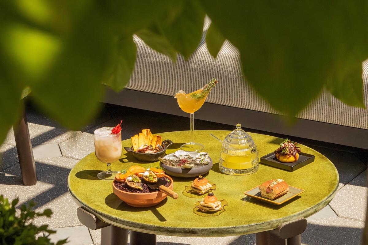 Outdoor round greenish table with a variety of tapas and cocktails, framed by leafy foreground plants on a sunny patio.
