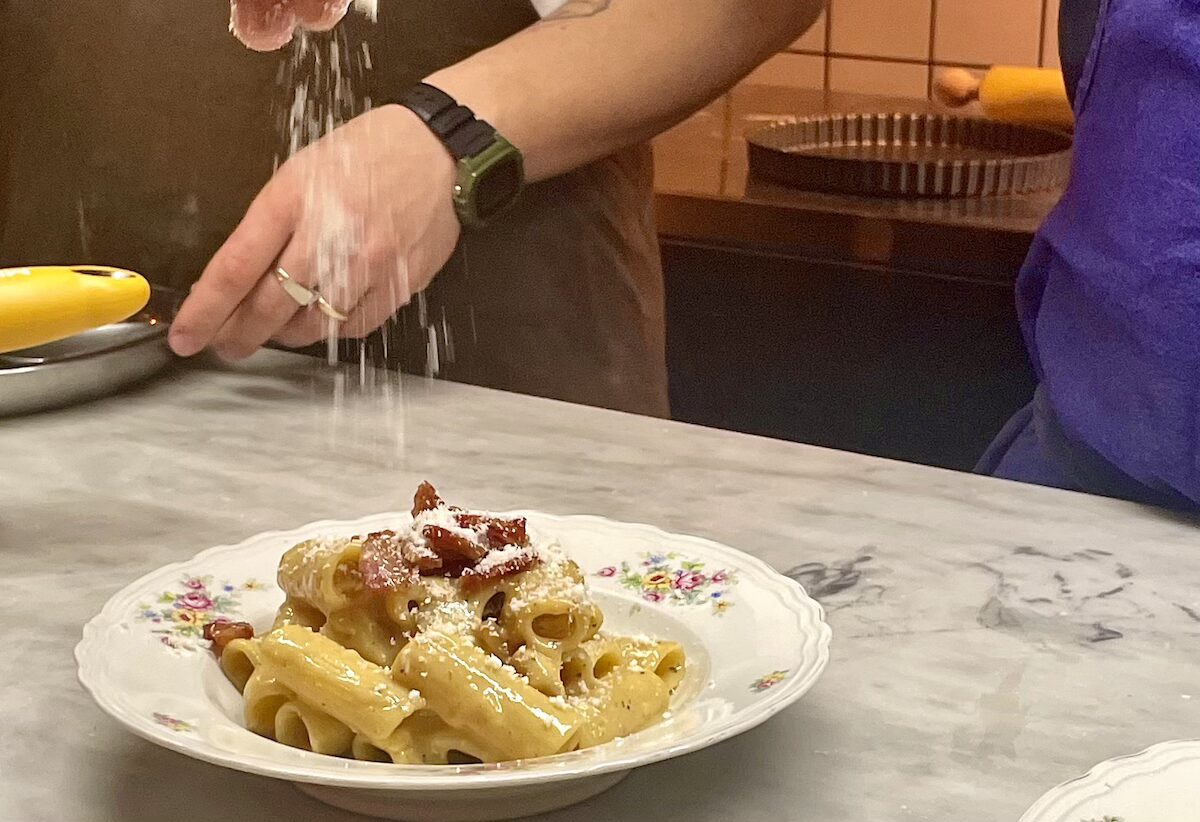 NY_RigatoniAllaCarbonara_DavidFarley - DiningOut Chef sprinkles grated cheese over a plate of creamy penne pasta on a marble counter.