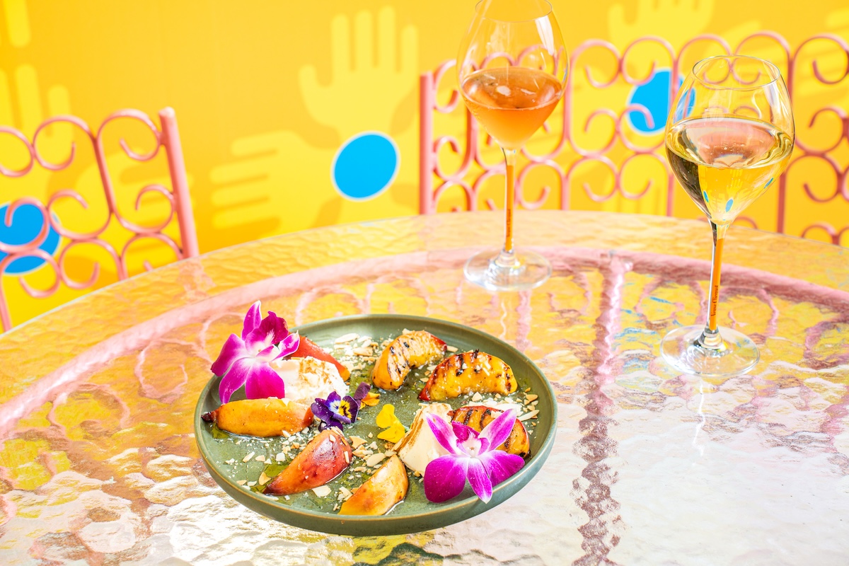 NY_StandardEastVillage_GrilledPeaches - DiningOut Green plate with a colorful tropical dish and purple orchids on a glass table, with two wine glasses and a bright yellow background.