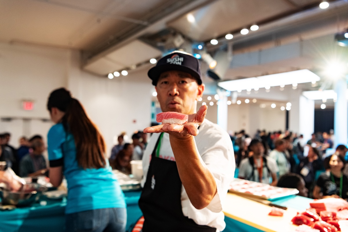 Sushi chef in a cap and apron extending a slice of tuna toward the camera while a crowded event runs in the background