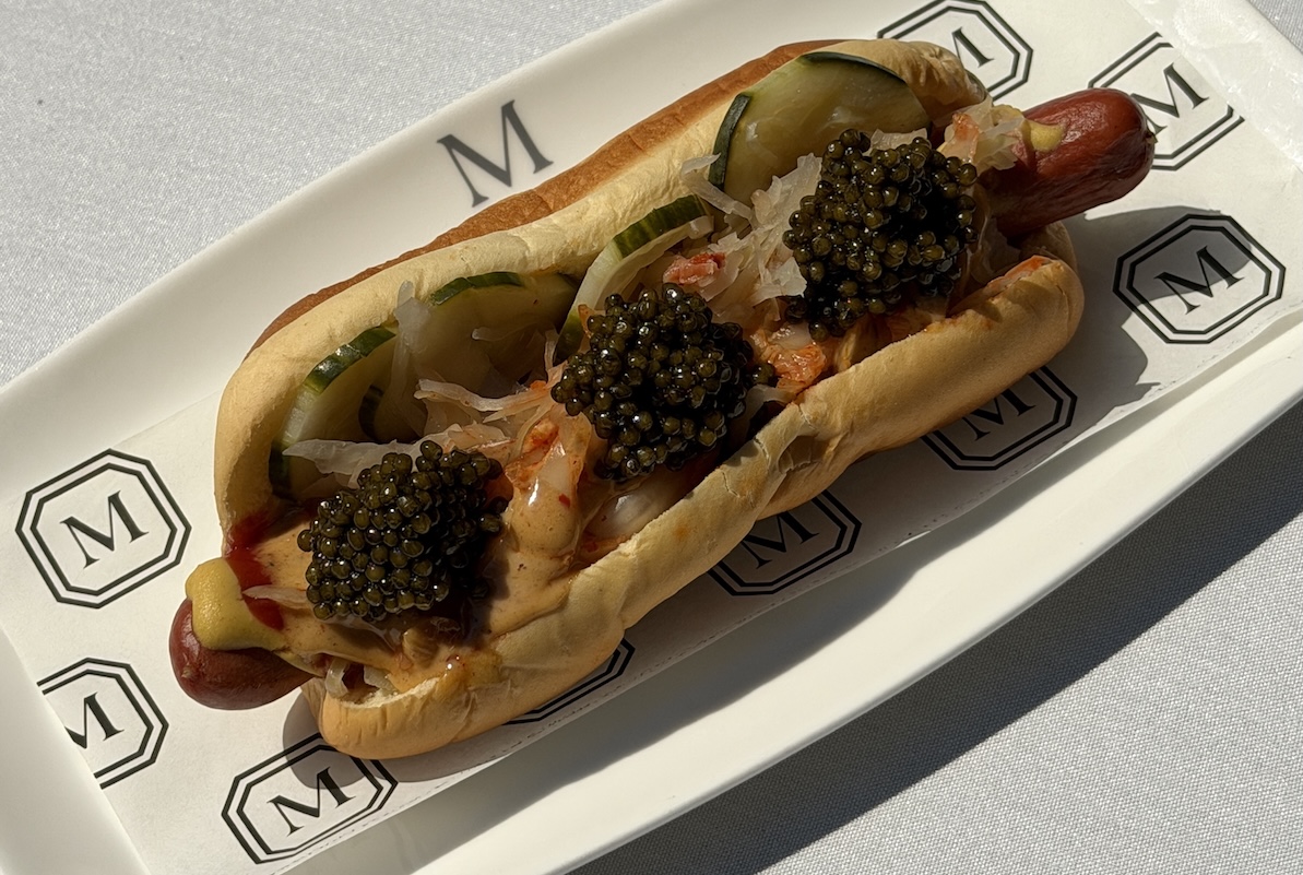 NY_TheMarkHotDog - DiningOut Deluxe hot dog in a toasted bun topped with black caviar, onions, pickles, and sauce on a branded white plate.