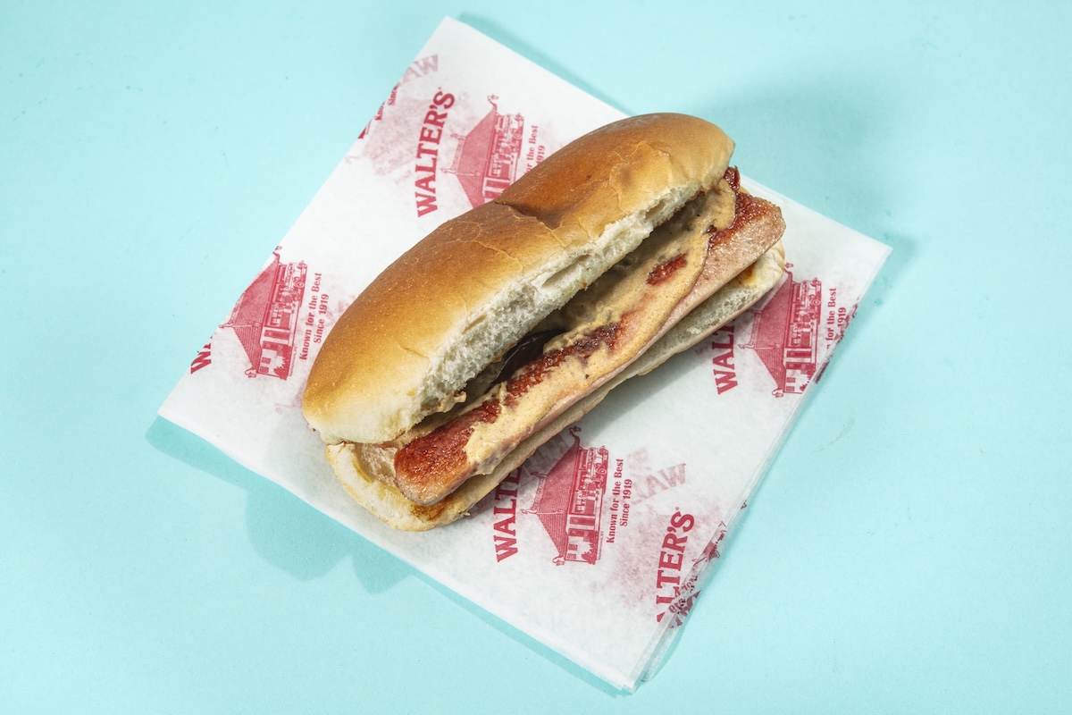 Hot dog in a toasted bun with toppings on Walther’s branded paper, set on a pastel blue background as a close-up snack photo.