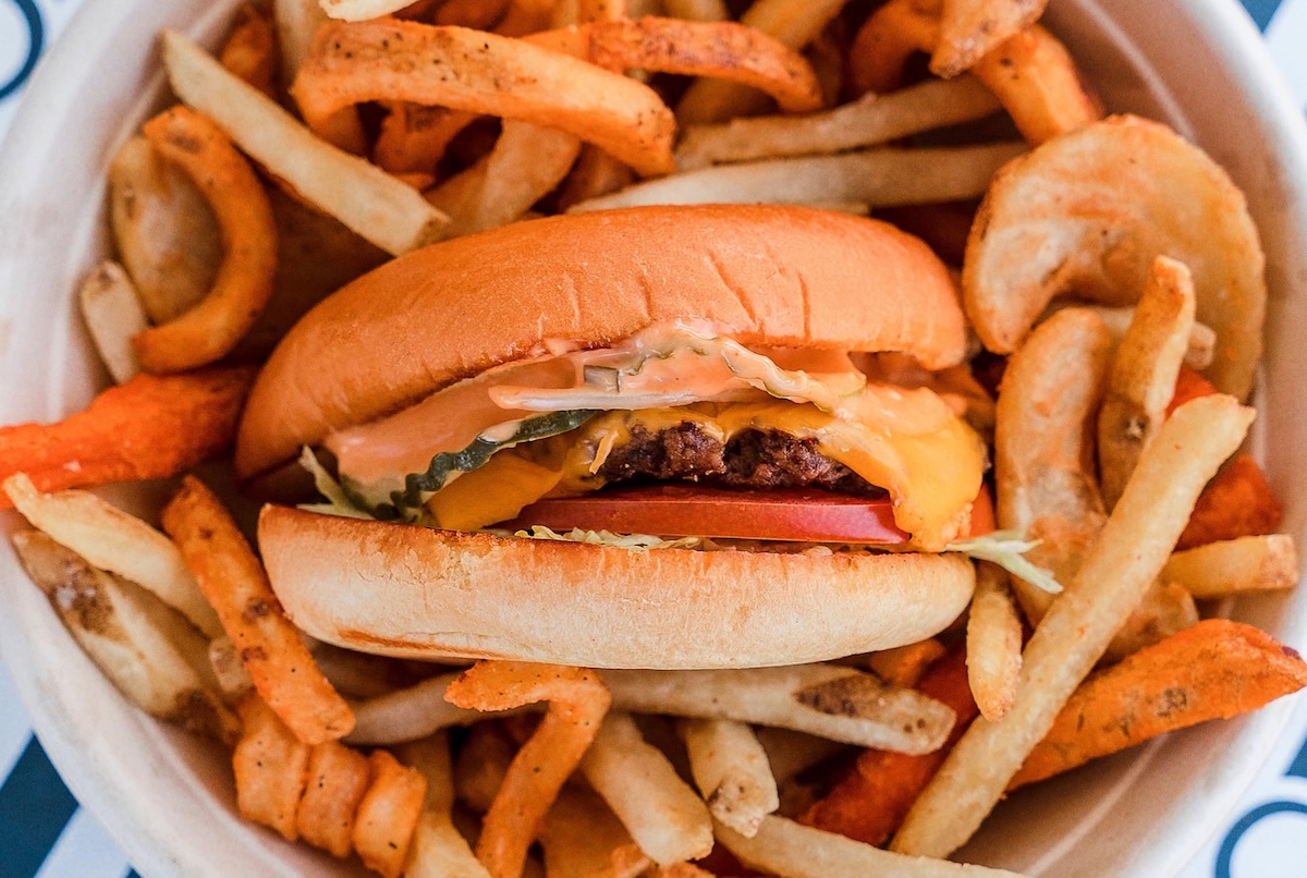 Cheeseburger with lettuce, tomato, pickles and melted cheese in a soft bun, surrounded by seasoned fries and onion rings in a bowl