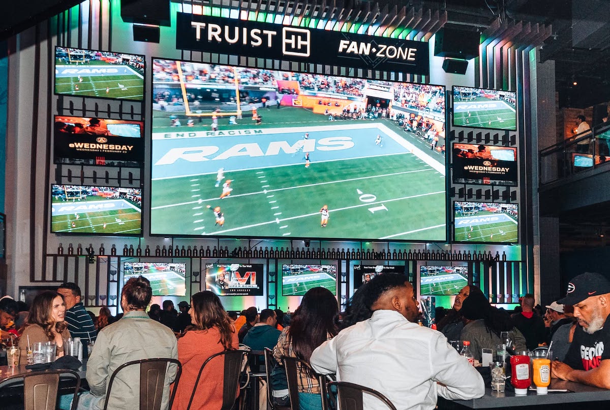 Busy sports bar with a large central TV showing an American football game; smaller screens around display the action and fans watch from tables and chairs.