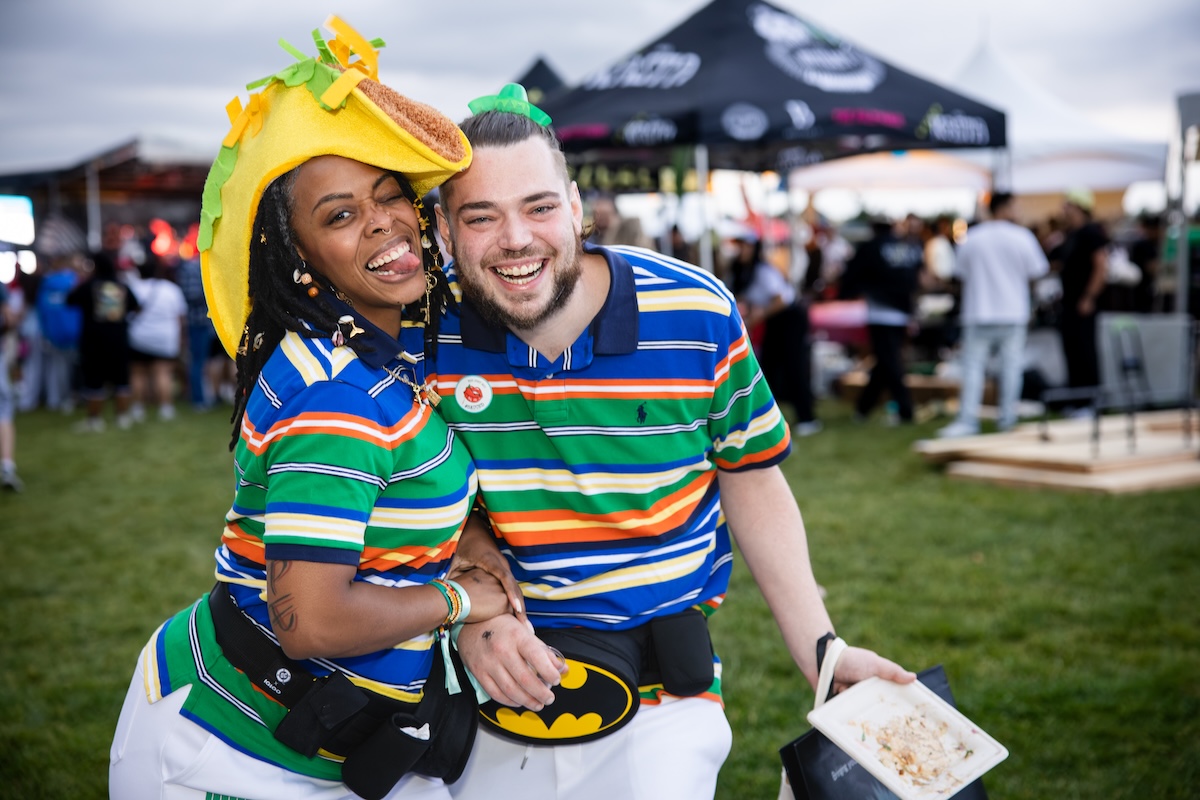 Two friends at an outdoor festival pose and smile for the camera; one wears a taco hat and colorful striped shirts.