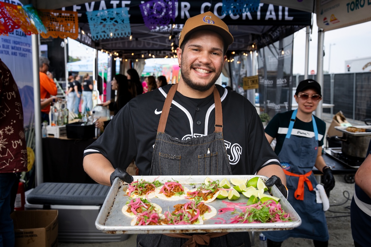 Smiling chef in a brown cap and denim apron holds a tray of loaded tacos with lime wedges at an outdoor food festival.