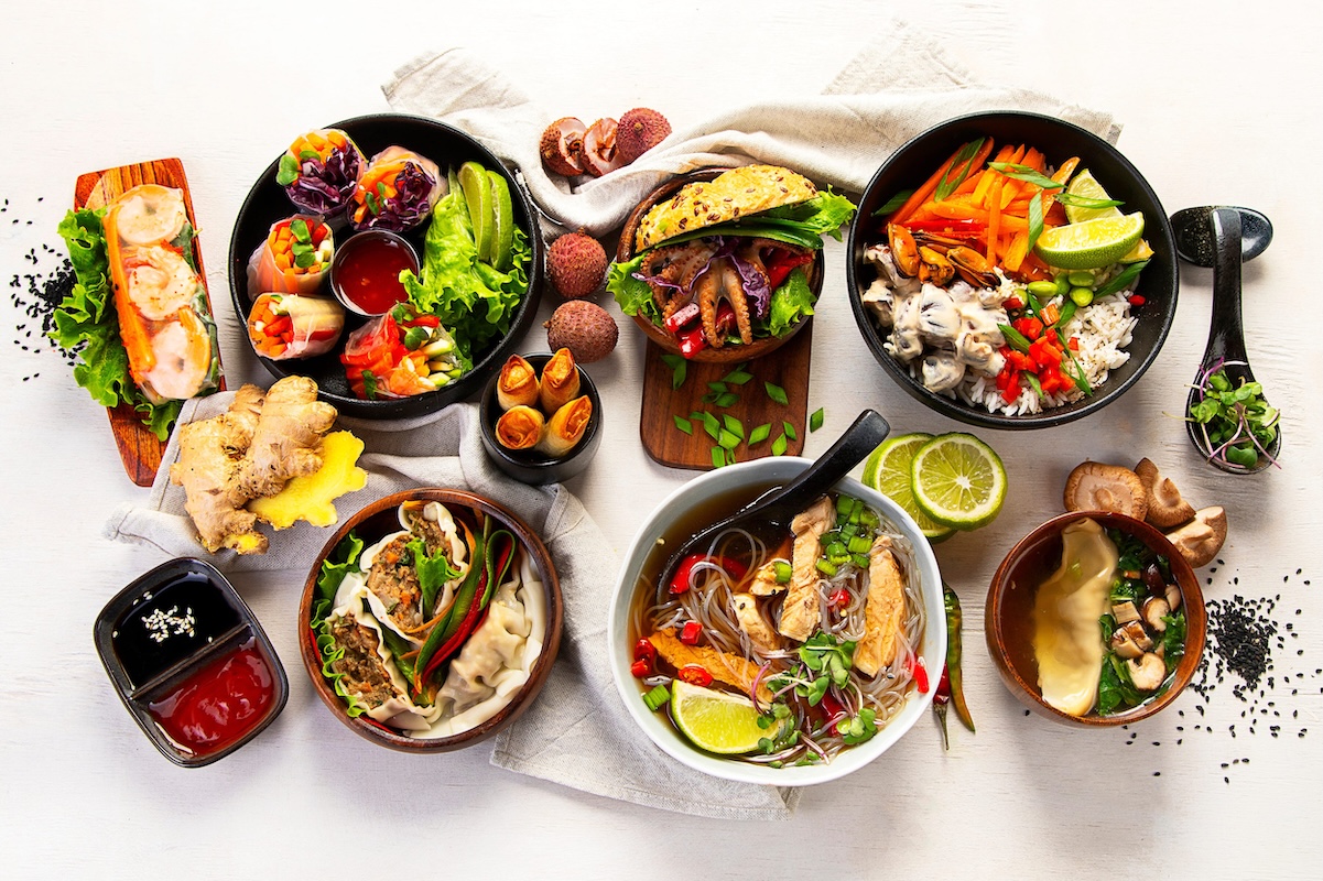 Top-down view of a colorful spread: sushi rolls, dumplings, wraps, ramen, and bowls with vegetables and lime wedges on a light table.