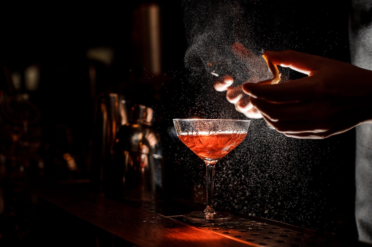 Bartender in black apron and blue shirt sprays an orange peel in cocktail glass with ice at a bar counter