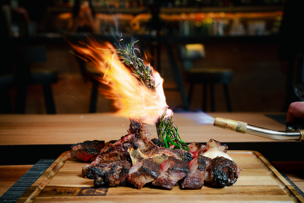 A long-bone tomahawk steak, dry-aged in Chr Steak's glass-walled aging room, is finished tableside with a flaming sprig of rosemary. | Photo by Southern Exposure Media Group