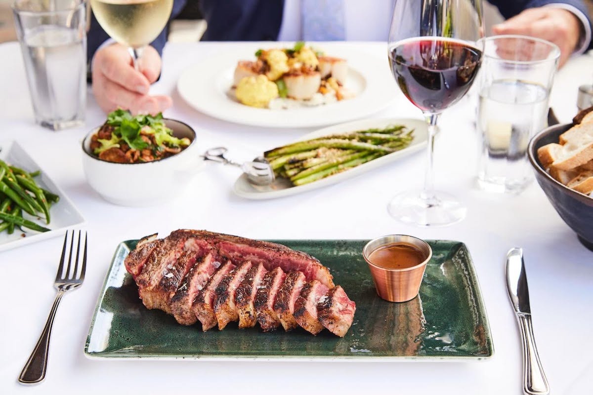 Al Biernat's DAL - DiningOut Sliced medium-rare steak on a green plate with sauce in a copper cup, surrounded by a formal dining table setup and side dishes.