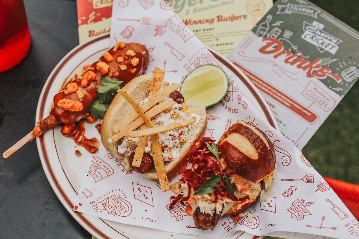 Plate with two seafood sliders: lobster roll topped with crispy tortilla strips and a side sausage-like skewer with crunchy toppings, lime wedge nearby.