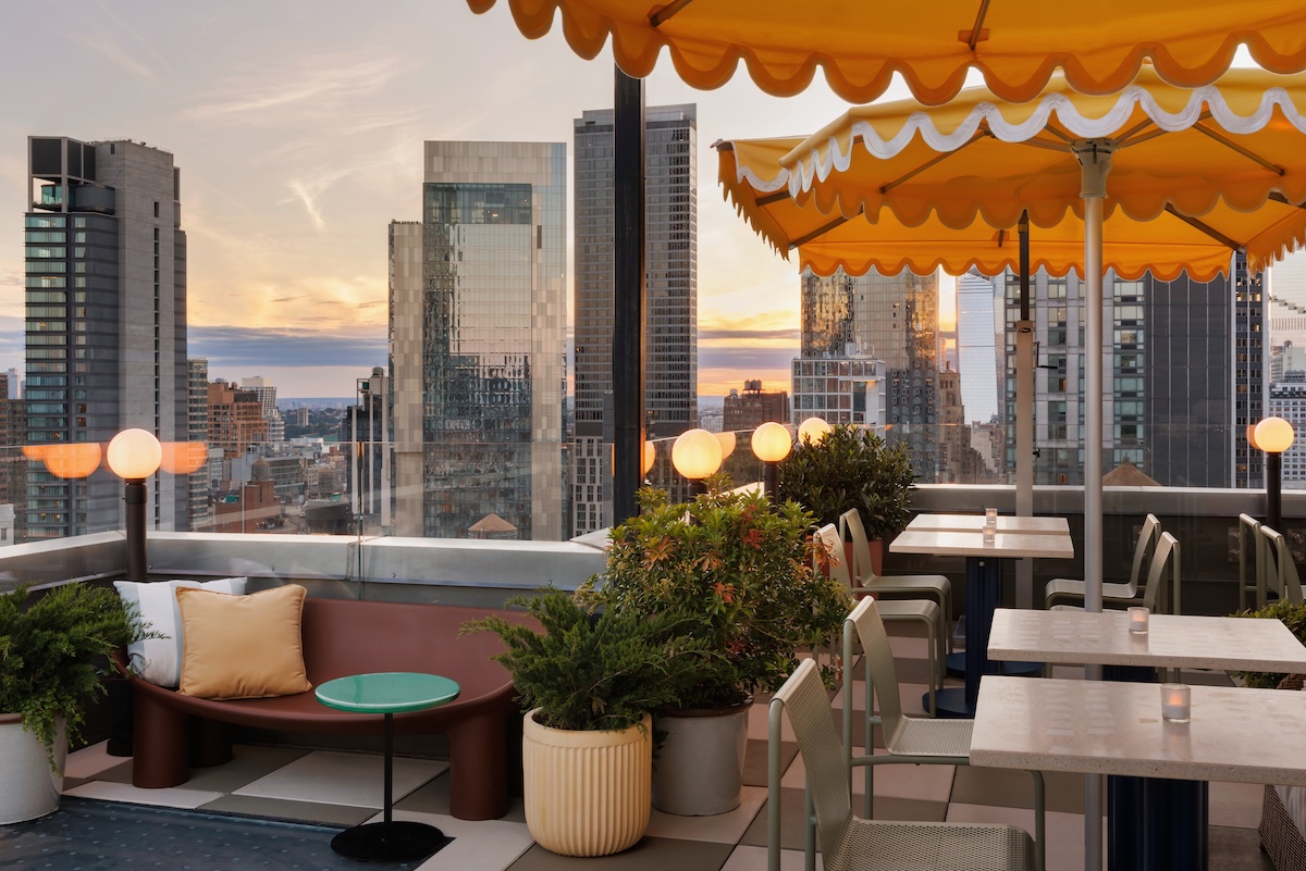 Rooftop cafe seating with yellow canopies and plants, overlooking a city skyline at sunset.